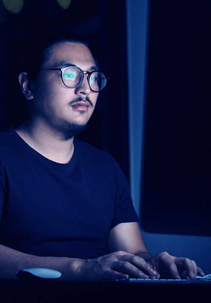 A man sits in the dark at their desk. They are typing on their computer.