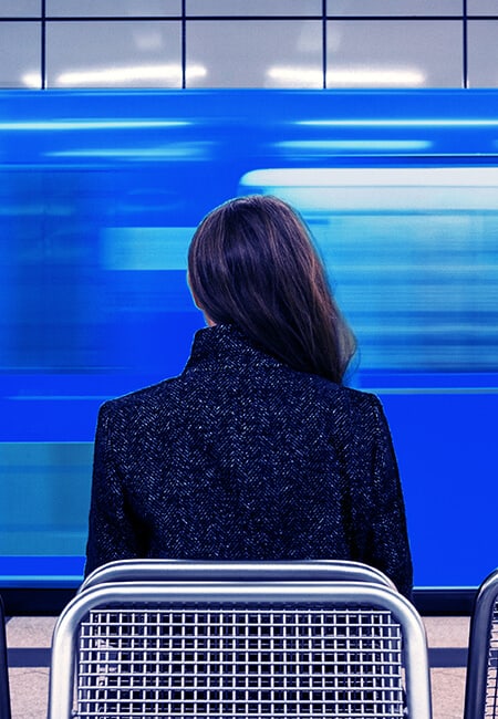 A woman sits in a train station, there is a blue train speeding past in a blur of motion.